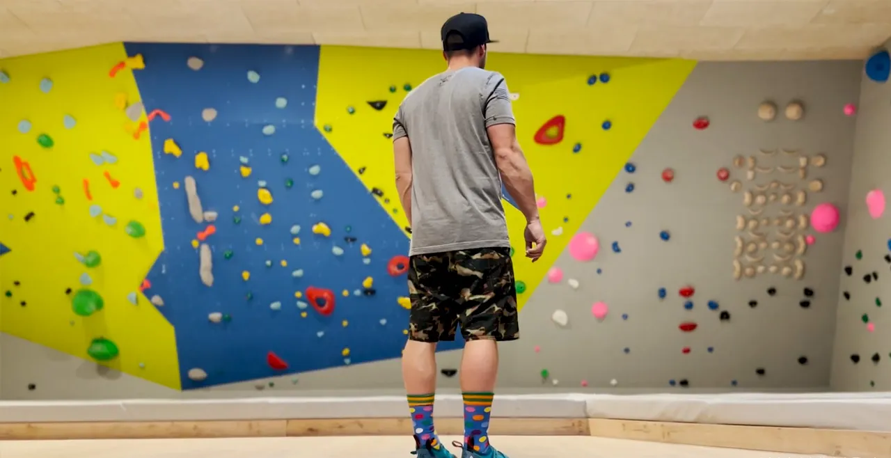 Ein junger Mann, welcher mit dem Rücken zur Kamera vor der Boulderwand steht.